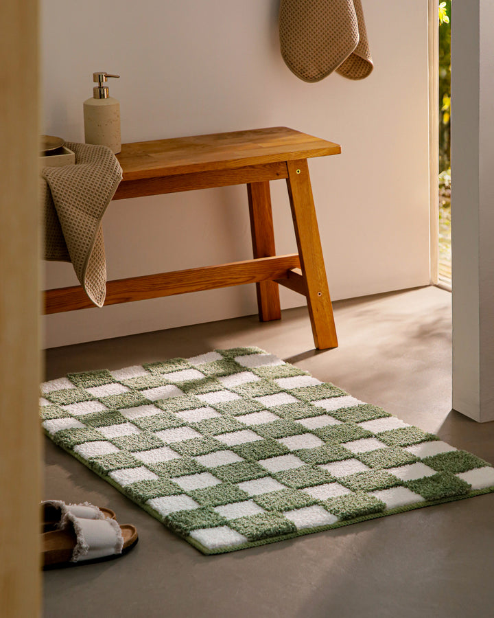 Badteppich Bobbi Grün im Badezimmer neben Holzbank auf hellem Boden liegend, rechteckige Matte mit Karomuster