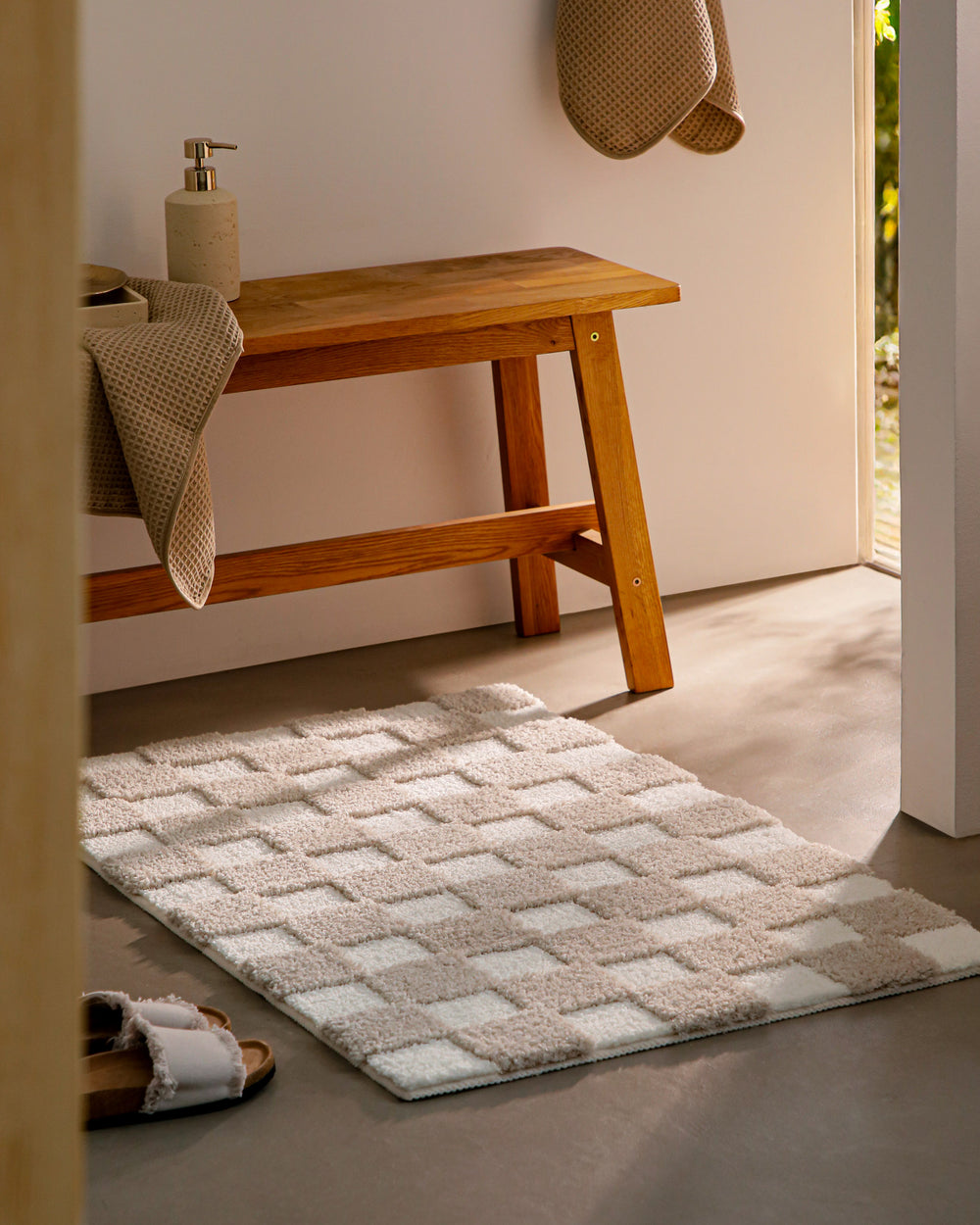 Badteppich Bobbi Beige im Badezimmer neben Holzbank auf hellem Boden liegend, rechteckige Matte mit Karomuster
