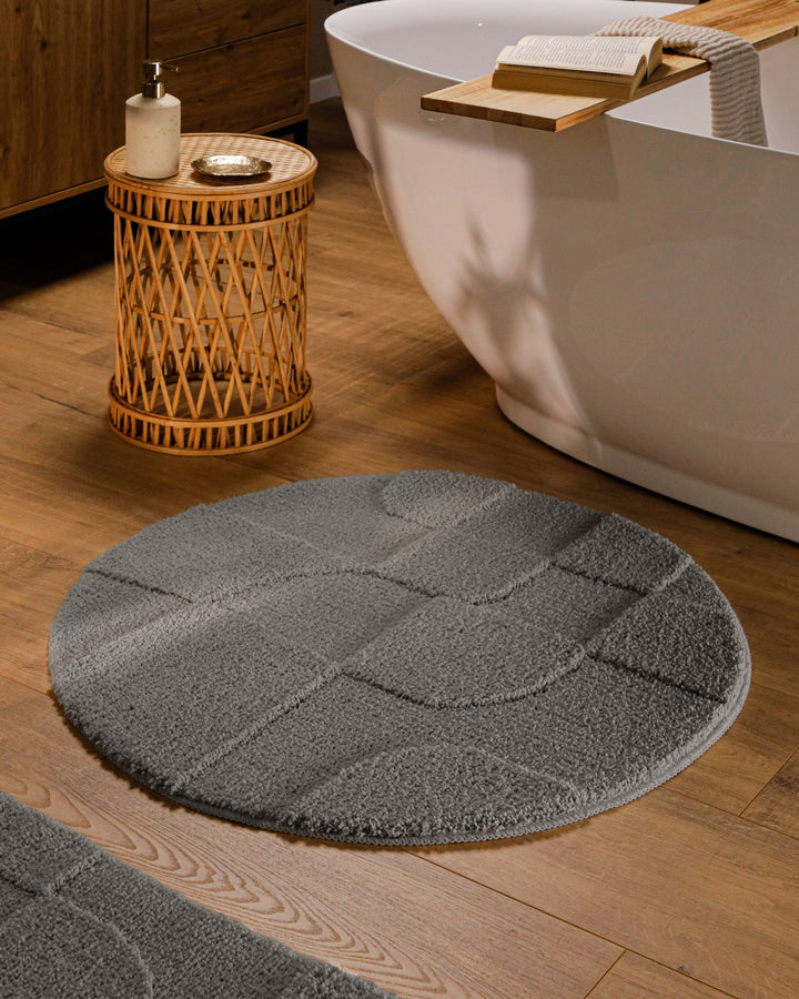Runder Badteppich Davie Basalt Grau im Badezimmer vor Badewanne auf Holzboden, strukturiertes Linienrelief im Flor sichtbar