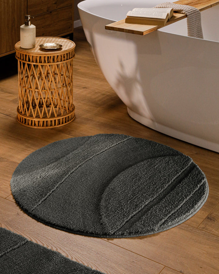 Runder Badteppich Marc Anthrazit Grau im Badezimmer vor Badewanne auf Holzboden, strukturiertes Linienrelief im Flor sichtbar