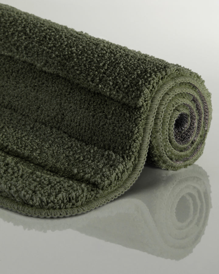 Badteppich Marc Kiefer Grün aufgerollt auf hellem Hintergrund, Seitenansicht zeigt Florhöhe und Teppichkante