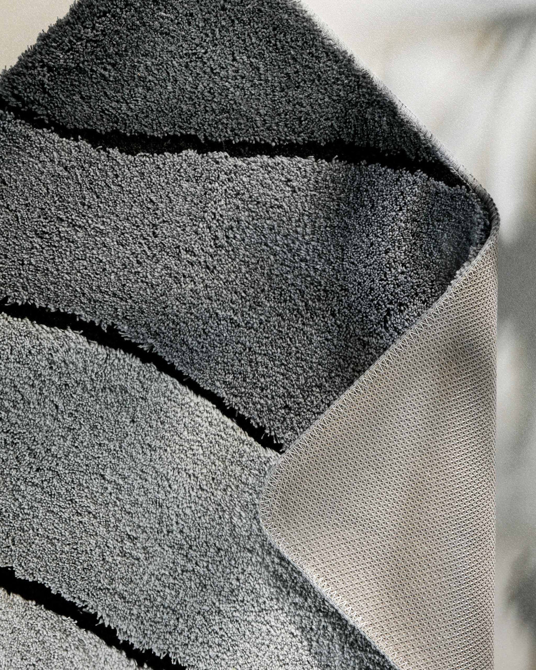 Nahaufnahme einer aufgeschlagenen Teppichkante – Flor in Dunkelgrau mit Wellendetail auf der Vorderseite und sichtbarem beigem Textilrücken