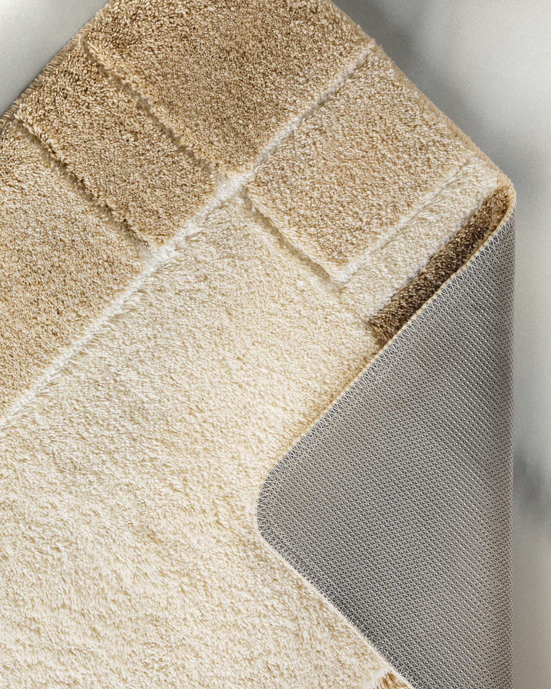 Nahaufnahme einer aufgeschlagenen Ecke des Badteppichs Villa Elba – Blockbordüre in Beige und Taupe auf der Vorderseite, darunter sichtbarer grauer Textilrücken