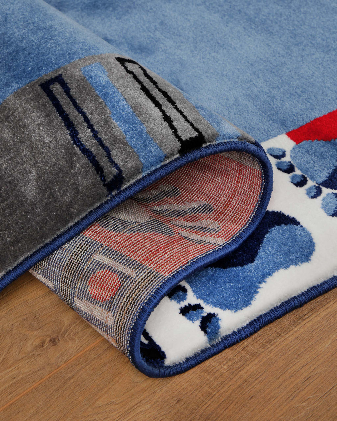 Kinderteppich Hand&Feet, umgeschlagene Ecke zeigt grauen Teppichrücken und blauen Rand mit Fußabdruck-Motiv auf Holzboden