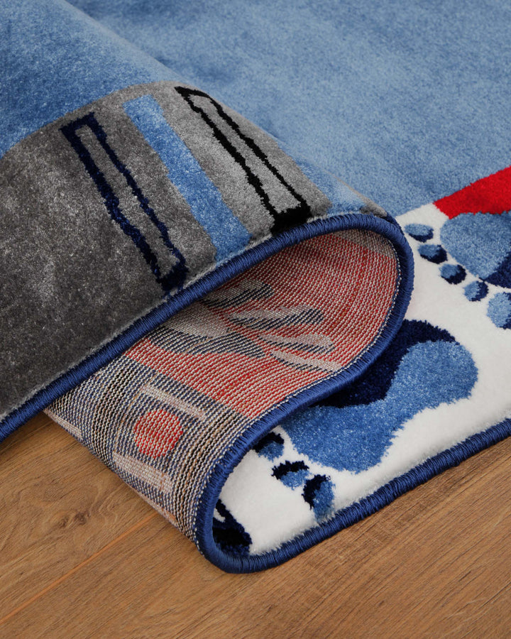 Kinderteppich Hand&Feet, umgeschlagene Ecke zeigt grauen Teppichrücken und blauen Rand mit Fußabdruck-Motiv auf Holzboden
