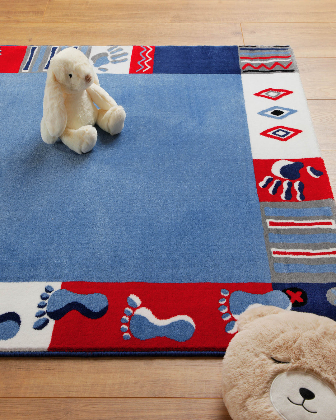 Ecke des Kinderteppichs Hand&Feet mit Teddybär als Deko, unifarbene blaue Mitte und gemusterter Rand mit Fußabdrücken
