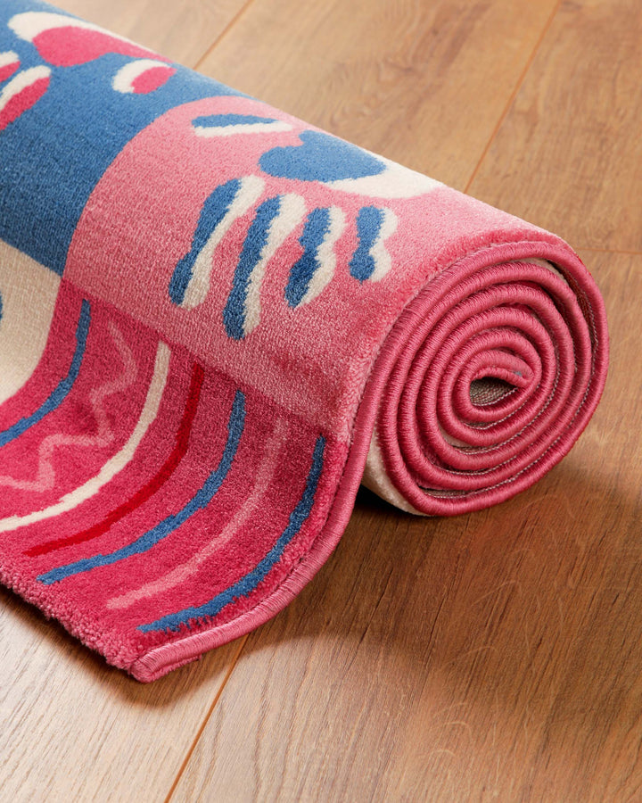 Zusammengerollter Kinderteppich Hand&Feet mit pinken Handabdrücken und blauen Streifen auf Holzboden
