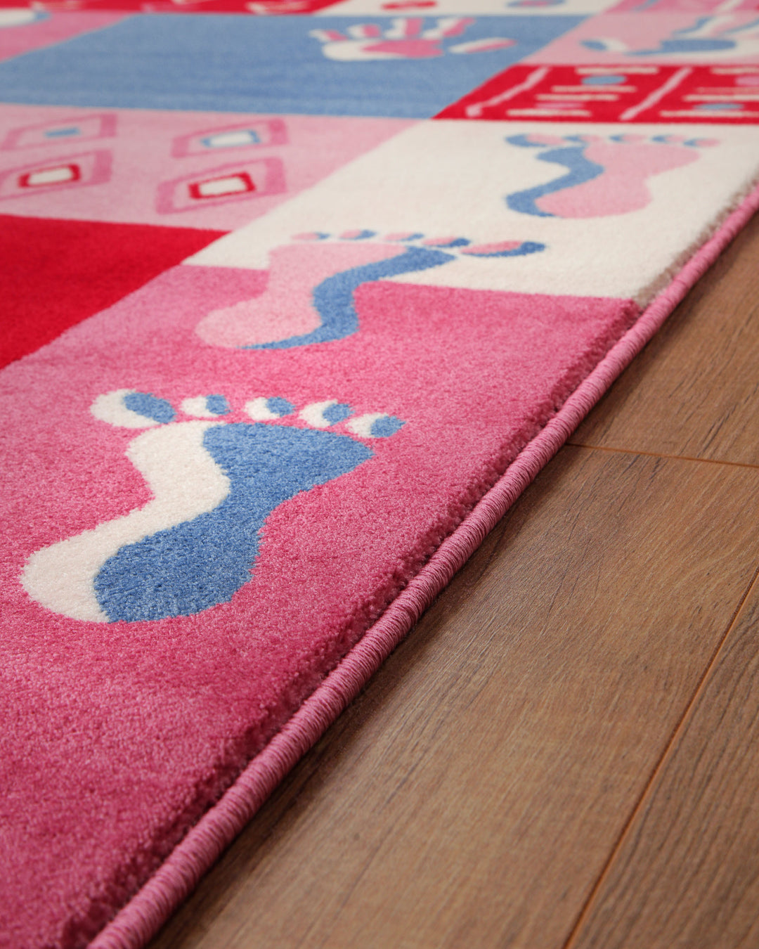Kante des Kinderteppichs Hand&Feet mit Fußabdruck-Muster in Pink, Blau und Weiß auf Holzdielenboden