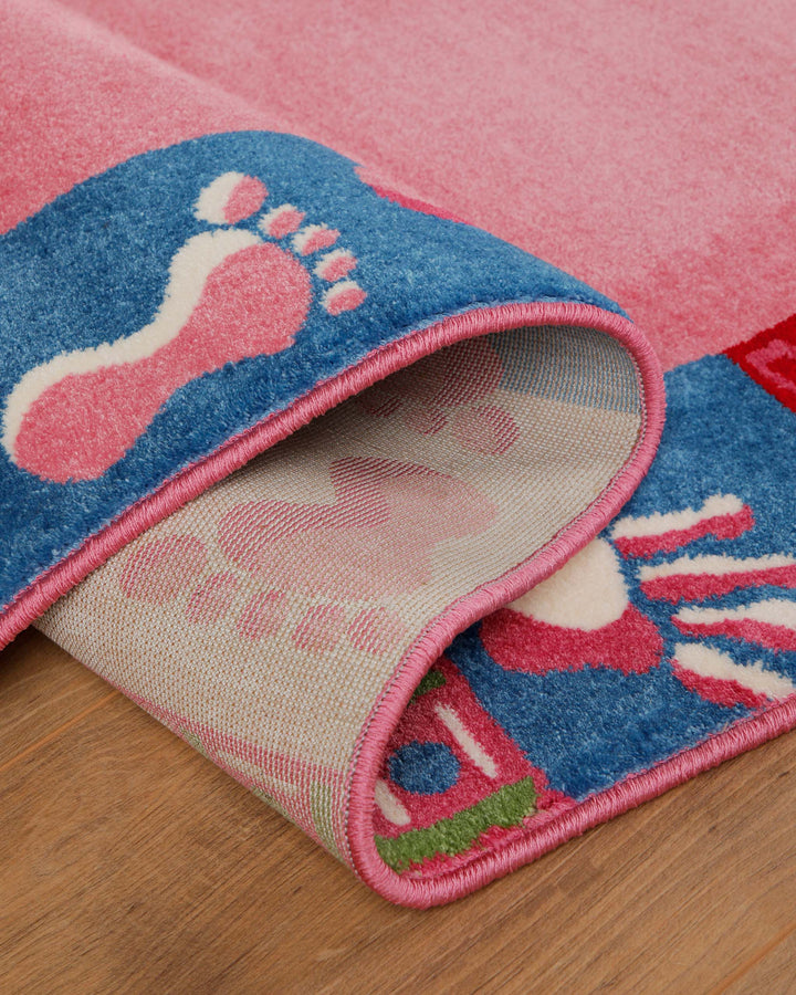 Kinderteppich Hand&Feet, umgeschlagene Ecke zeigt Teppichrücken und pinken Rand mit Fußabdruck-Motiv auf Holzboden