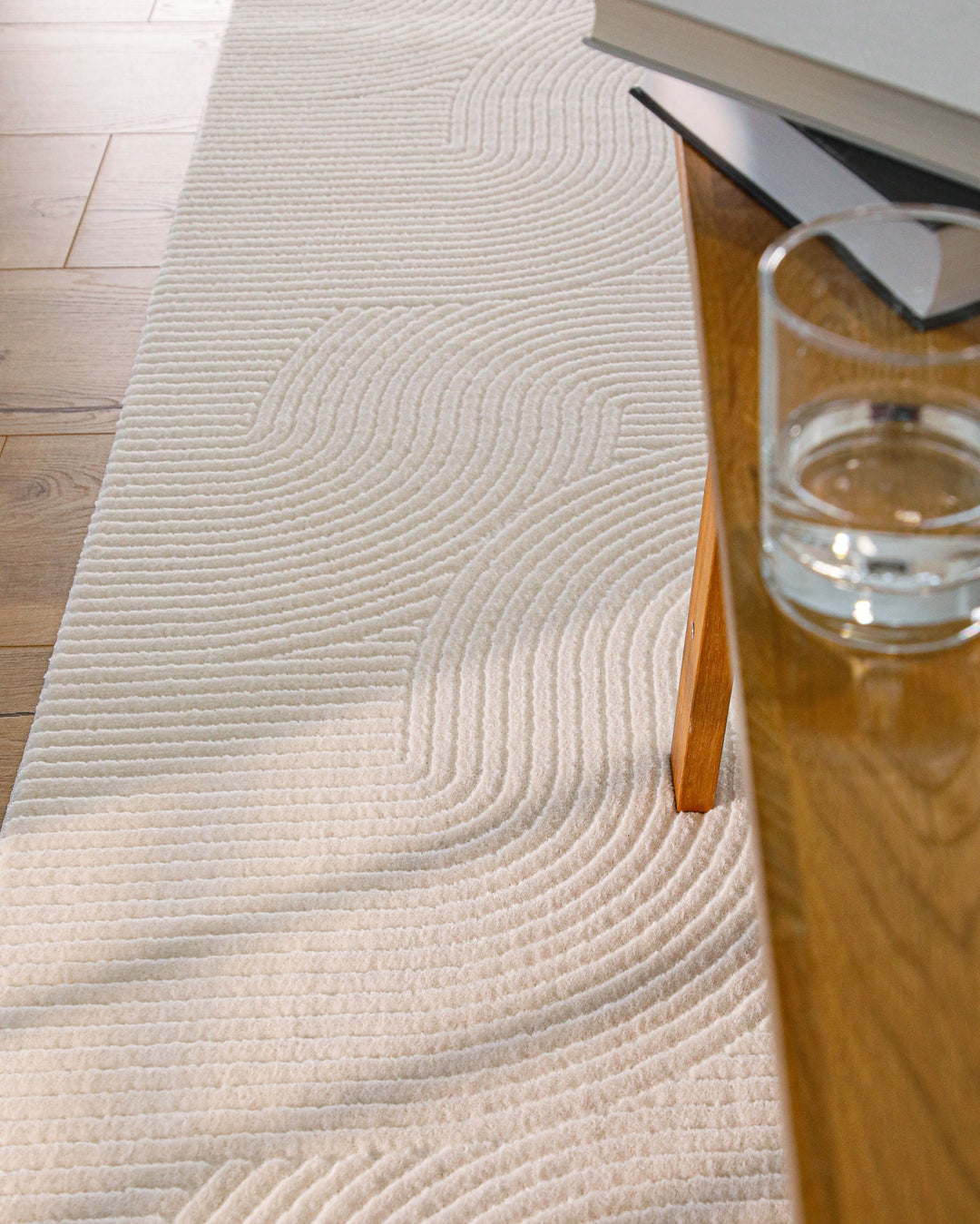 Nuuk Arc Teppich creme-weiß unter Holztisch mit Glas und Büchern